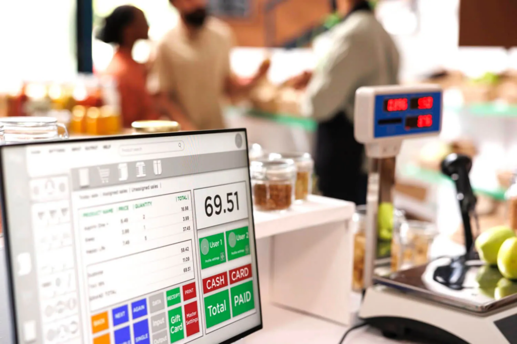 A digital Point of Sale (POS) terminal screen displaying a grocery transaction total, with blurred store customers in the background.