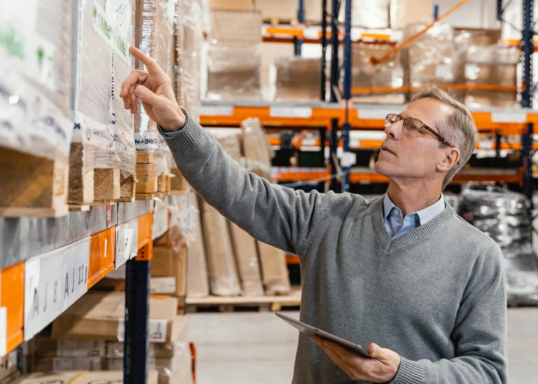 A supply chain manager inspecting inventory on tall warehouse shelving while holding a digital tablet.