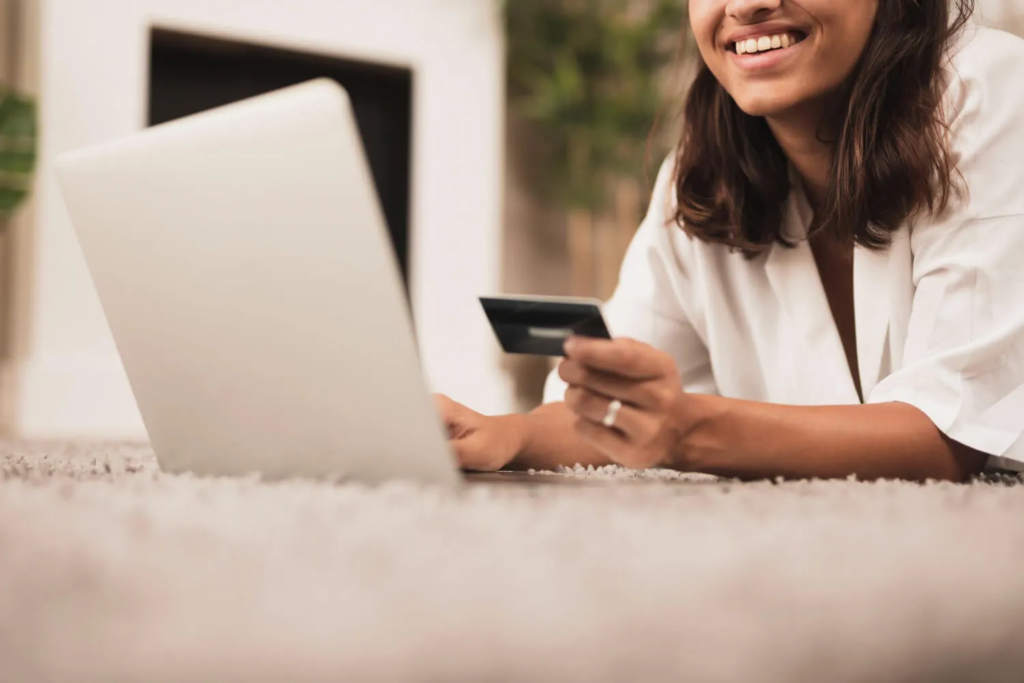 A smiling woman lying on the floor while using a laptop and holding a credit card to shop online.