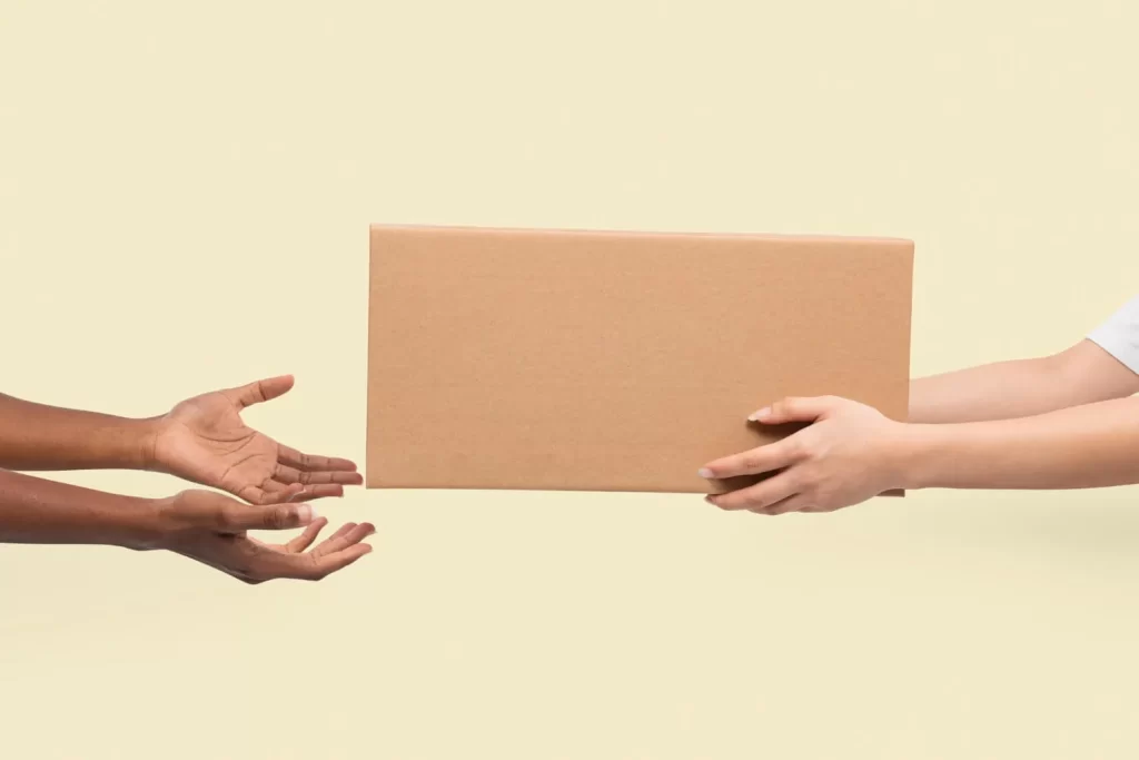 A close-up of a product package being handed from one person to another against a neutral studio background.