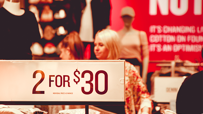 A retail store promotional sign displaying a "2 FOR $30" deal with shoppers browsing clothing in the blurred background.