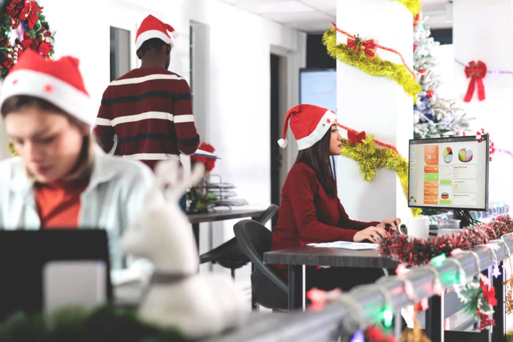 Office employees wearing Santa hats and working at computers in a workplace decorated with Christmas trees and holiday ornaments.